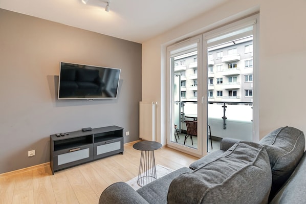 A modern living room with a flat-screen TV, grey sofa, and a sliding glass door leading to a balcony. - Bogucice Modern Apartment (Katowice)