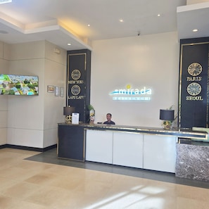 A hotel reception desk with a digital clock displaying 'New York' and 'Paris' on the wall, a flat-screen TV mounted on the wall, and a receptionist behind the desk. - New Lux. 2 BR Condo 63 SQM - 2 Pools, Gym,  woow seaviews, Beach 5-6Pax (Lapu-Lapu City)