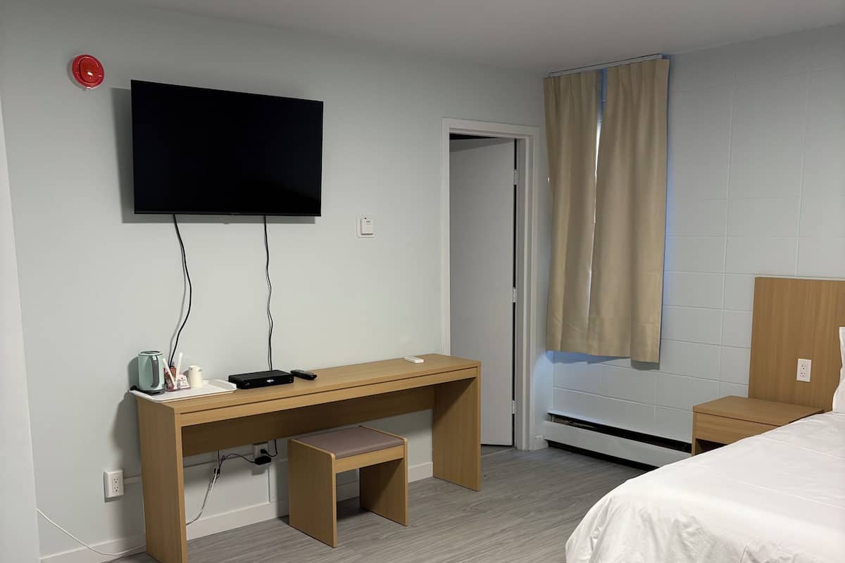 A hotel room with a bed, a desk with a chair, a television mounted on the wall, and a window with curtains.
