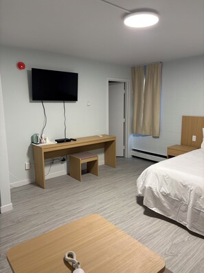 Quarto de hotel com cama, escrivaninha com cadeira, televisão na parede e janela com cortinas.