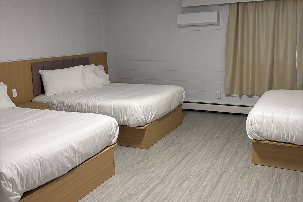 Habitación de hotel con tres camas, una lámpara en el techo, un equipo de aire acondicionado y una ventana con cortinas.