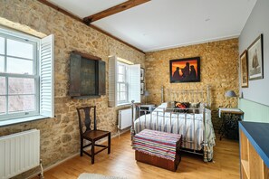 Quarto com paredes de pedra, uma cama com cabeceira de metal, uma cadeira, uma mesa e um quadro na parede.