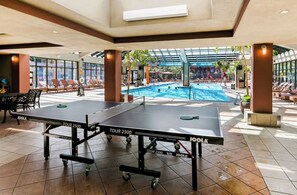 Dos mesas de ping pong JoolA en un espacio interior iluminado por el sol, con piscina y sillas de descanso.