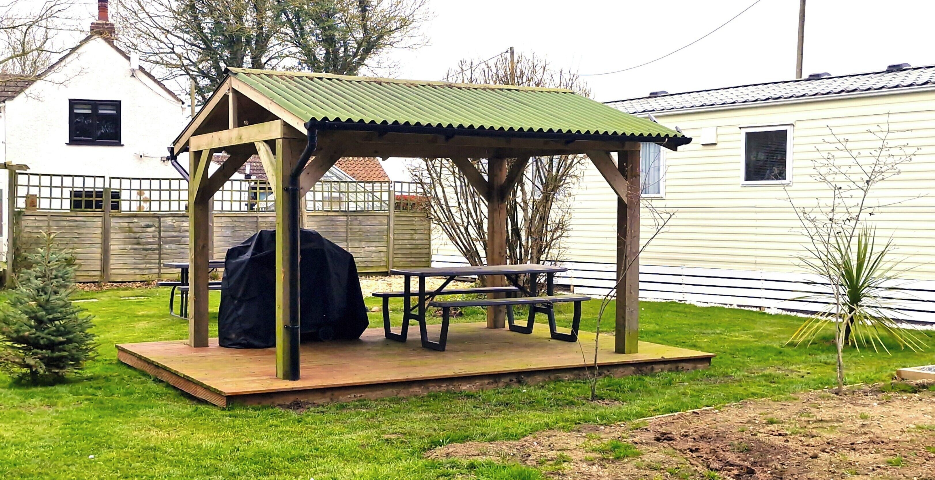 A wooden gazebo with a metal roof, a picnic table, and a barbecue grill in a backyard.