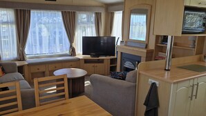 Interior compacto de uma motorhome, com área de estar, televisão, pequena mesa e cozinha compacta.