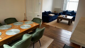 En matplats med ett träbord och gröna stolar, ett vardagsrum med en blå soffa och ett soffbord samt en vit dörr.
