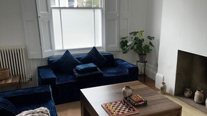 Ett vardagsrum med en soffa, ett soffbord med ett schackbräde, en öppen spis och utsikt över byggnader genom ett fönster.