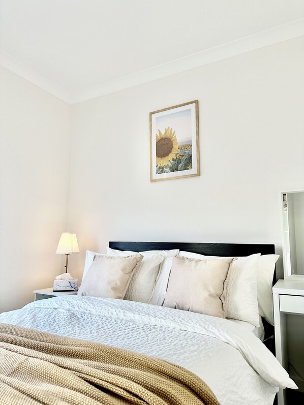 Una cama bien hecha con un edredón estampado, una lámpara de noche y un cuadro con una girasol en la pared.