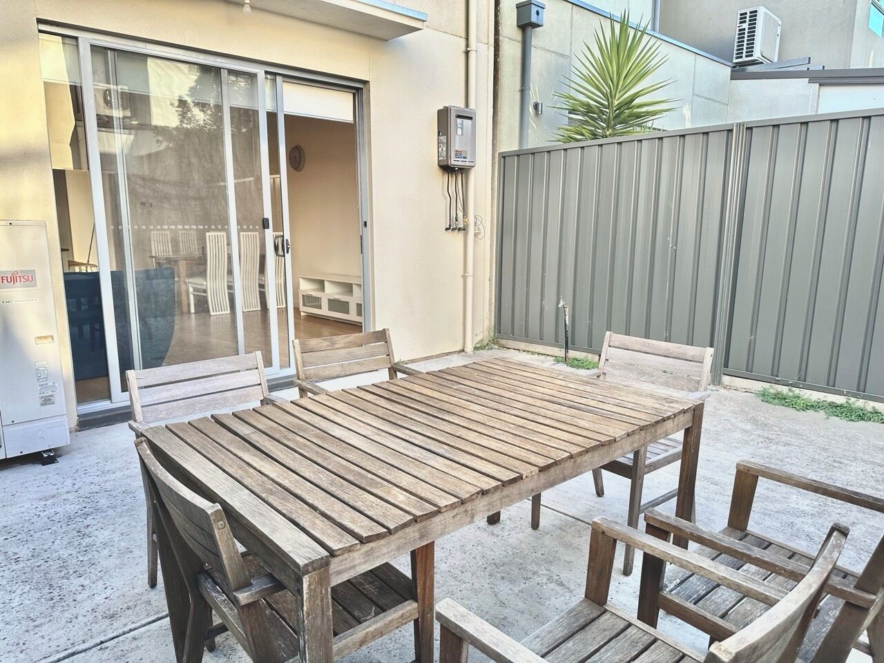 An outdoor patio with a wooden table and chairs, a sliding glass door, and a grey fence.