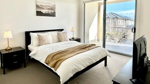 A bedroom with a bed, two bedside tables with lamps, a TV, a painting on the wall, and a balcony with a view.