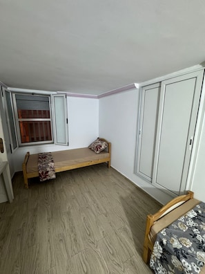 Un piset petit, d'una sola habitació, amb terra de fusta, un llit amb una colcha floral, una finestra amb persianes i un armari.