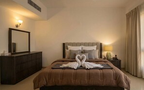 Une chambre d’hôtel avec un grand lit, une commode, un miroir, une table de chevet avec une lampe et une fenêtre avec des rideaux.