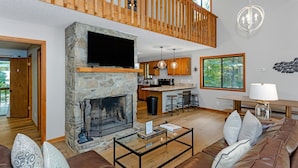 Un amplio salón con chimenea de piedra, un televisor montado encima y techo de madera, con vistas al exterior a través de un ventanal grande.