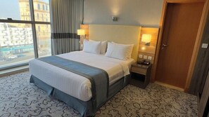 A hotel room with a large bed, bedside lamps, a wooden door, and a view of buildings through a window. - AL Shahba AL Faiha Hotel (Jeddah)