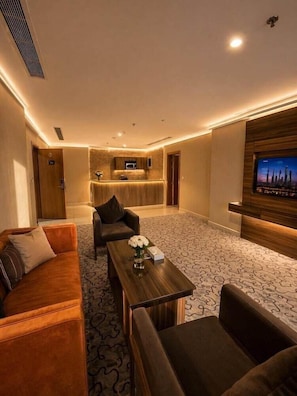 Habitación de hotel con sofá, mesa de centro, televisión, kitchenette y alfombra estampada.