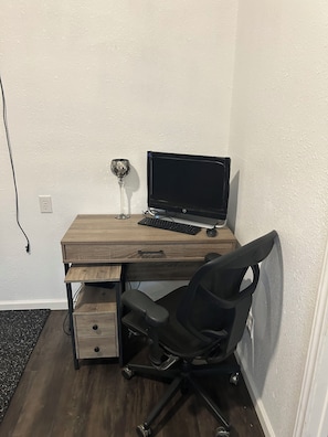 A corner workspace with a wooden desk, a black office chair, a computer monitor, keyboard, and a decorative glass vase.