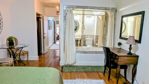 Quarto com cama, escrivaninha, cadeira, pequena mesa com planta, espelho e banheiro com banheira e vaso sanitário.