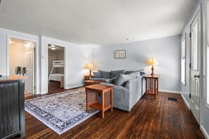 A room with a grey sofa, two wooden side tables with lamps, a patterned rug, a wooden cabinet, and a bunk bed in the background.