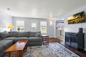 A living room with a sectional sofa, a wooden coffee table, a patterned rug, a kitchen area with a view, and a mounted TV.
