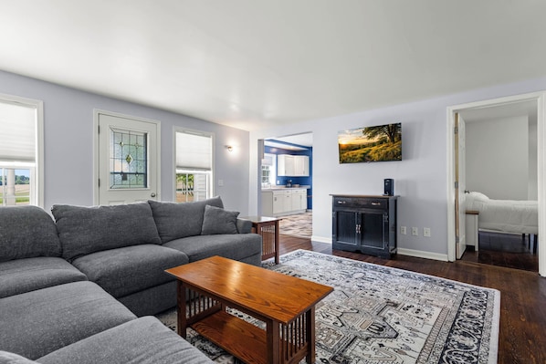 A living room with a sectional sofa, a wooden coffee table, a patterned rug, a kitchen in the background, and a painting on the wall.