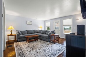A living room with a sectional sofa, two side tables with lamps, a coffee table, a patterned rug, a television, and a window with blinds.