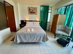 Quarto de hotel com cama, duas luminárias de cabeceira, cortina verde, sofá estampado e mesa de centro de vidro.