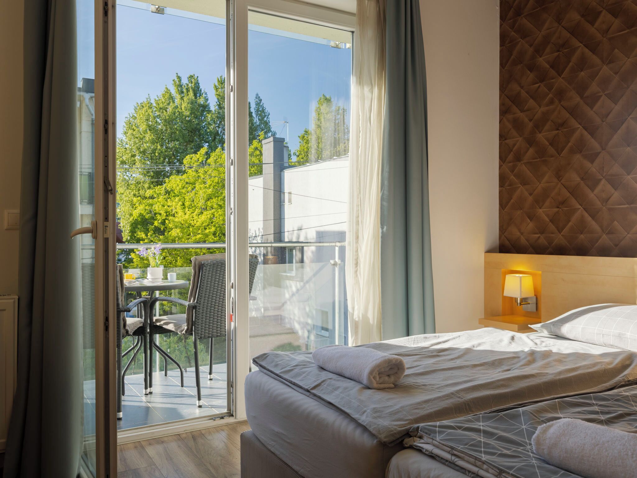 Une chambre d’hôtel avec un lit, un balcon doté d’une table et de chaises, et une vue sur les arbres et les bâtiments.