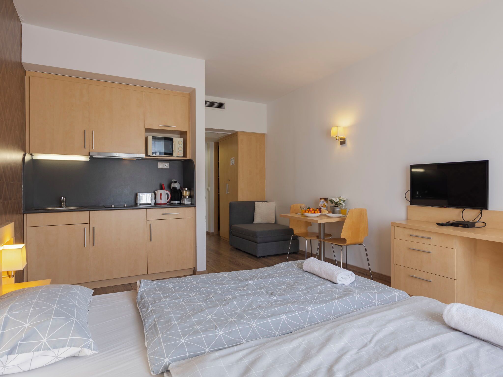 A modern hotel room with a kitchenette, a dining area, a TV, and a bed with geometric-patterned bedding.
