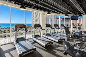 Ein Fitnessstudio mit Laufbändern und Ergometer, große Fenster mit Blick auf das Meer und Gebäude.