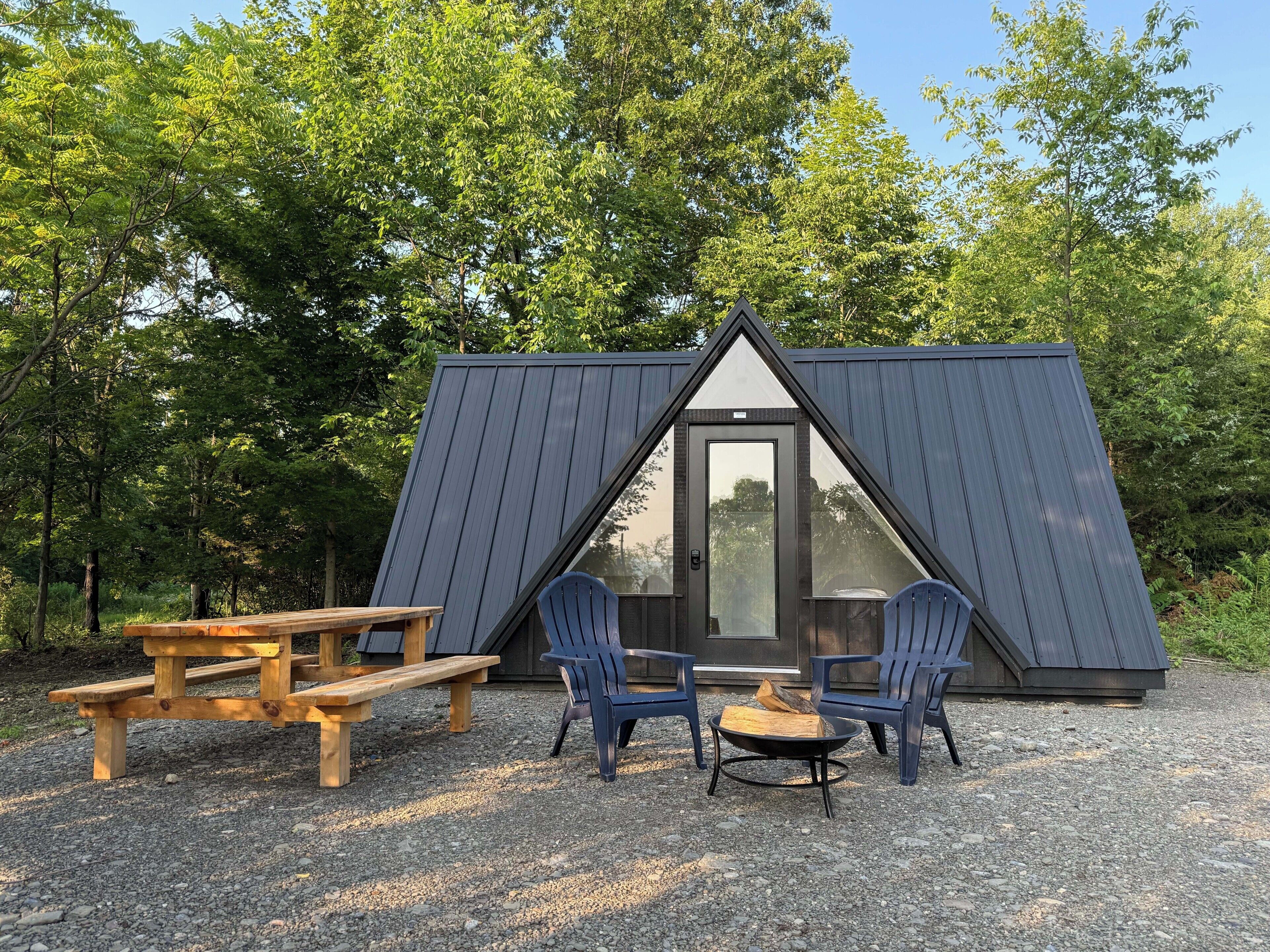 Una pequeña cabaña de madera triangular con puerta y ventanas, rodeada de árboles.