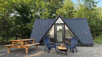Una pequeña cabaña de madera triangular con puerta y ventanas, rodeada de árboles.
