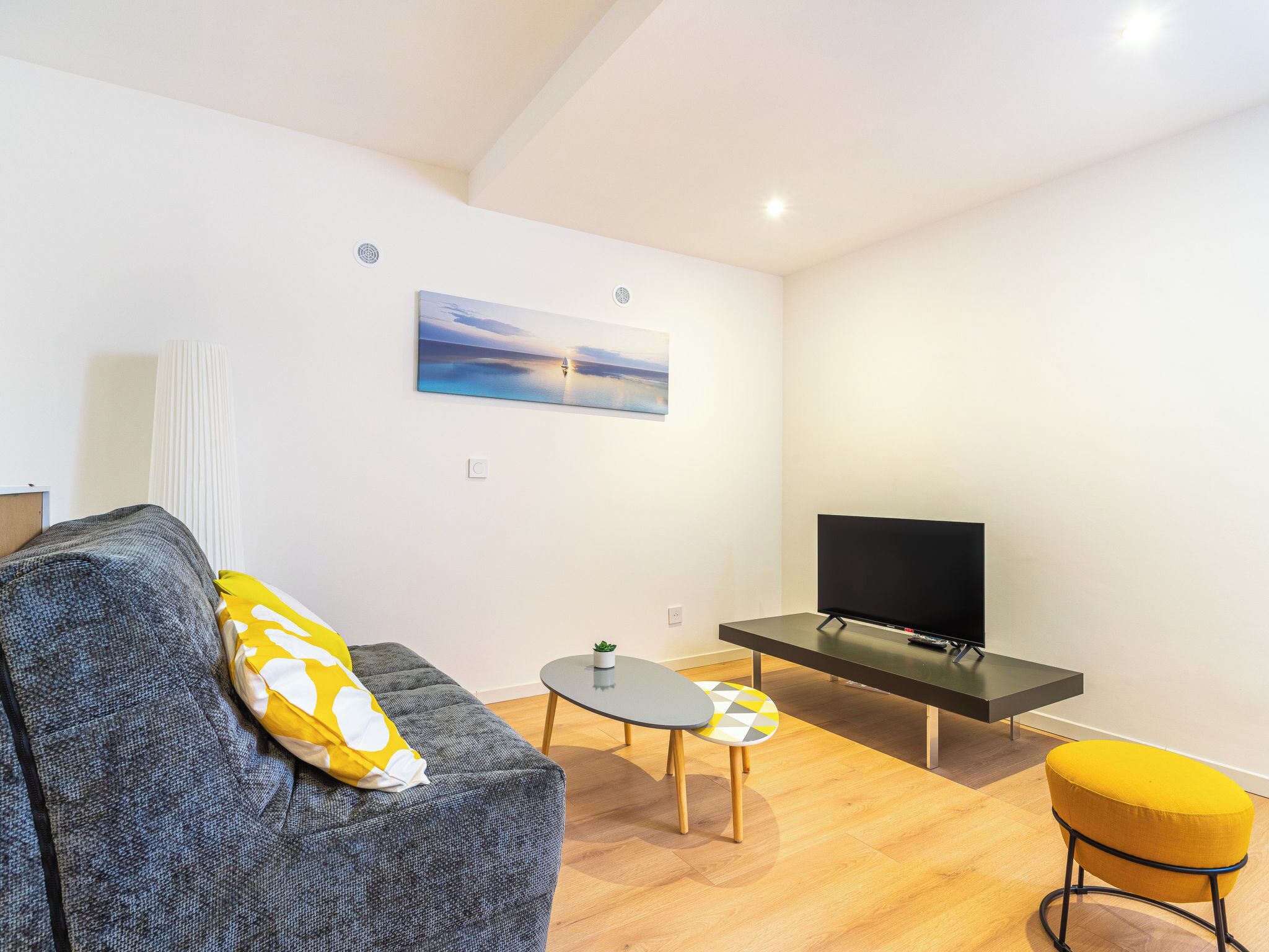 A modern living room with a sofa, a small table, a yellow ottoman, a flat-screen TV, and a painting on the wall.