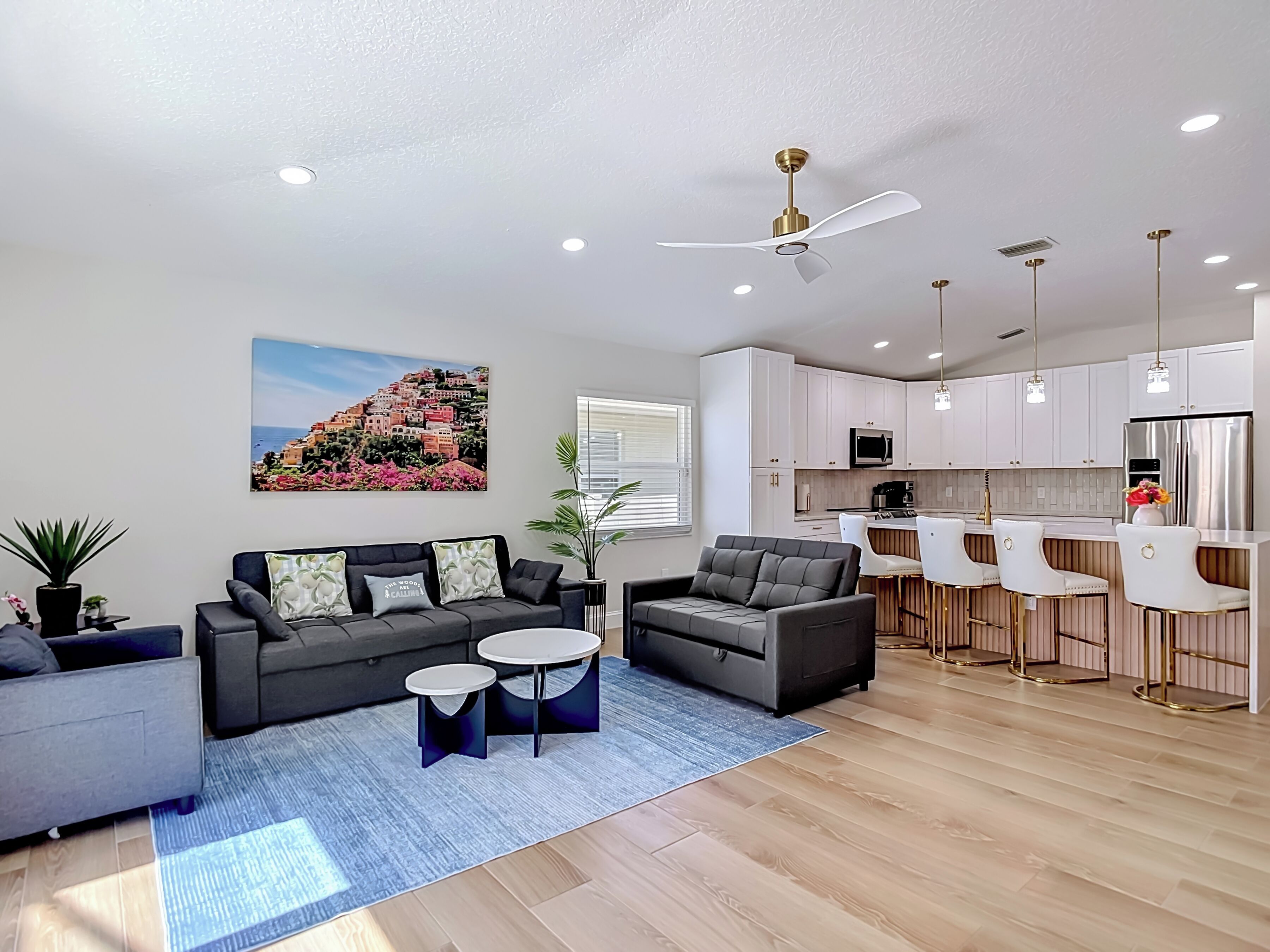 Una sala moderna con un sofá, dos sillones, una mesa de centro y una cocina con armarios blancos y un refrigerador de acero inoxidable.