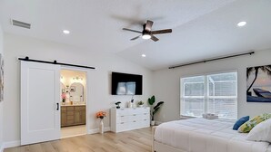 A bedroom with a bed, a ceiling fan, a window with blinds, a dresser, and a door leading to a bathroom. - Luxury Pool home, 1 mile from Bush Gardens (Tampa)