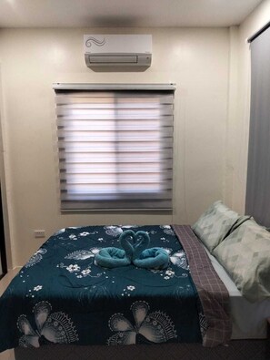A bedroom with a bed, a window with blinds, a wall-mounted air conditioner, and a decorative towel arrangement on the bed.