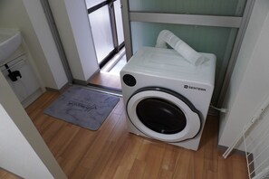 A compact washer and dryer unit with a white exterior, placed in a small laundry room with wooden flooring and a gray mat. - Within walking distance of Shibuya, Harajuku and Shinjuku! 86 sqm spacious family size apartment with balcony (渋谷区)