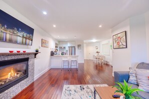 A modern living room with a fireplace, a kitchen in the background, and a dining area.