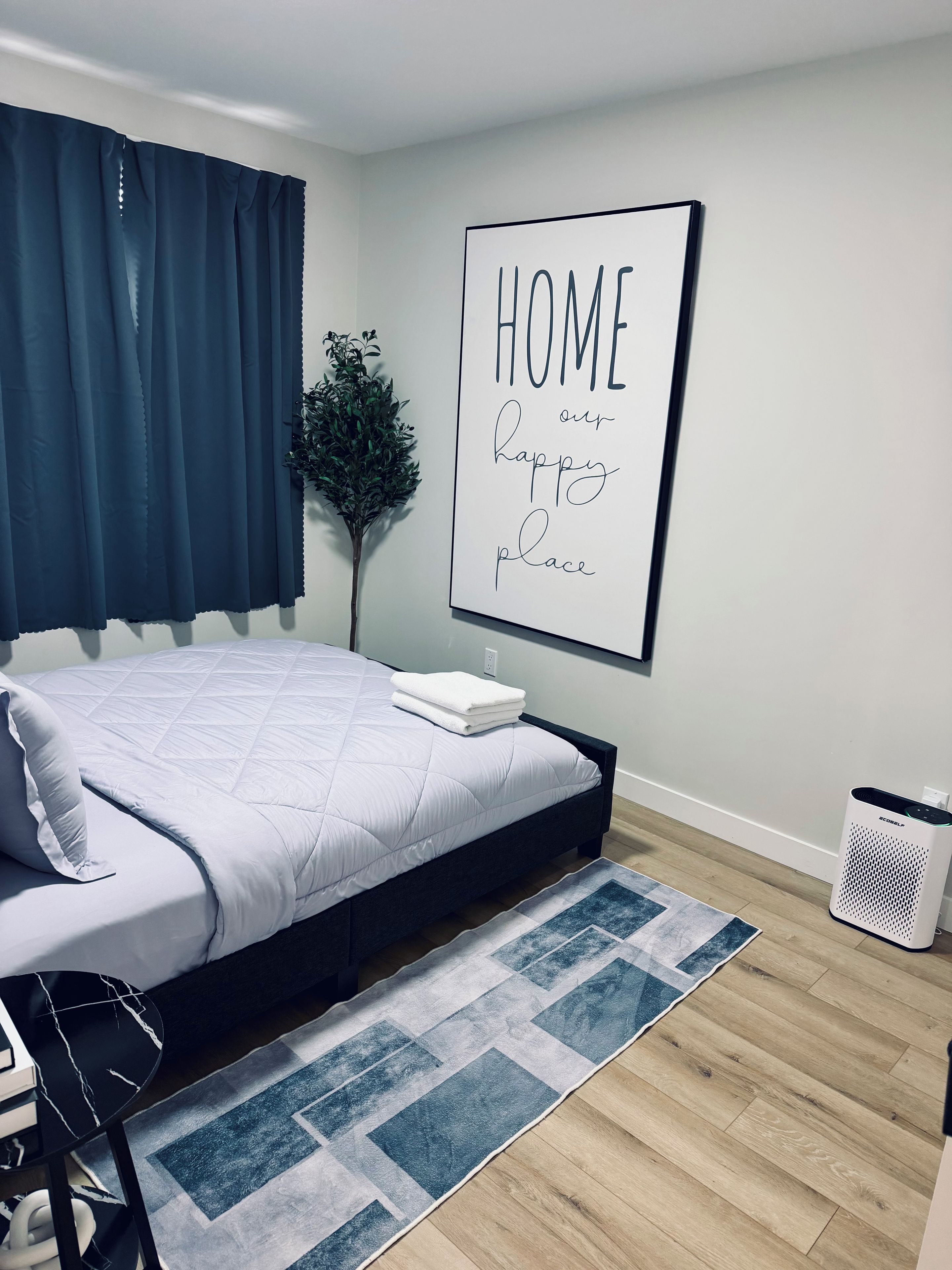 A neatly made bed with a white comforter and pillows, a blue and white patterned rug, a framed sign on the wall, and a small potted plant.