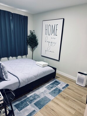 Una cama bien hecha con edredón blanco y almohadas, una alfombra estampada en azul y blanco, un cartel enmarcado en la pared y una pequeña planta en maceta.