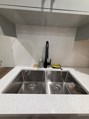 A kitchen with two stainless steel sinks, a faucet, a soap dispenser, a sponge, and a yellow cloth. - The Onyx Suite: Minimalist Luxury w/ Gym & Parking (Regina)