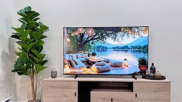 A modern living room with a flat-screen TV, a wooden cabinet, a potted plant, a glass table, and a book with 'OH, HAPPY DAY' on the cover.