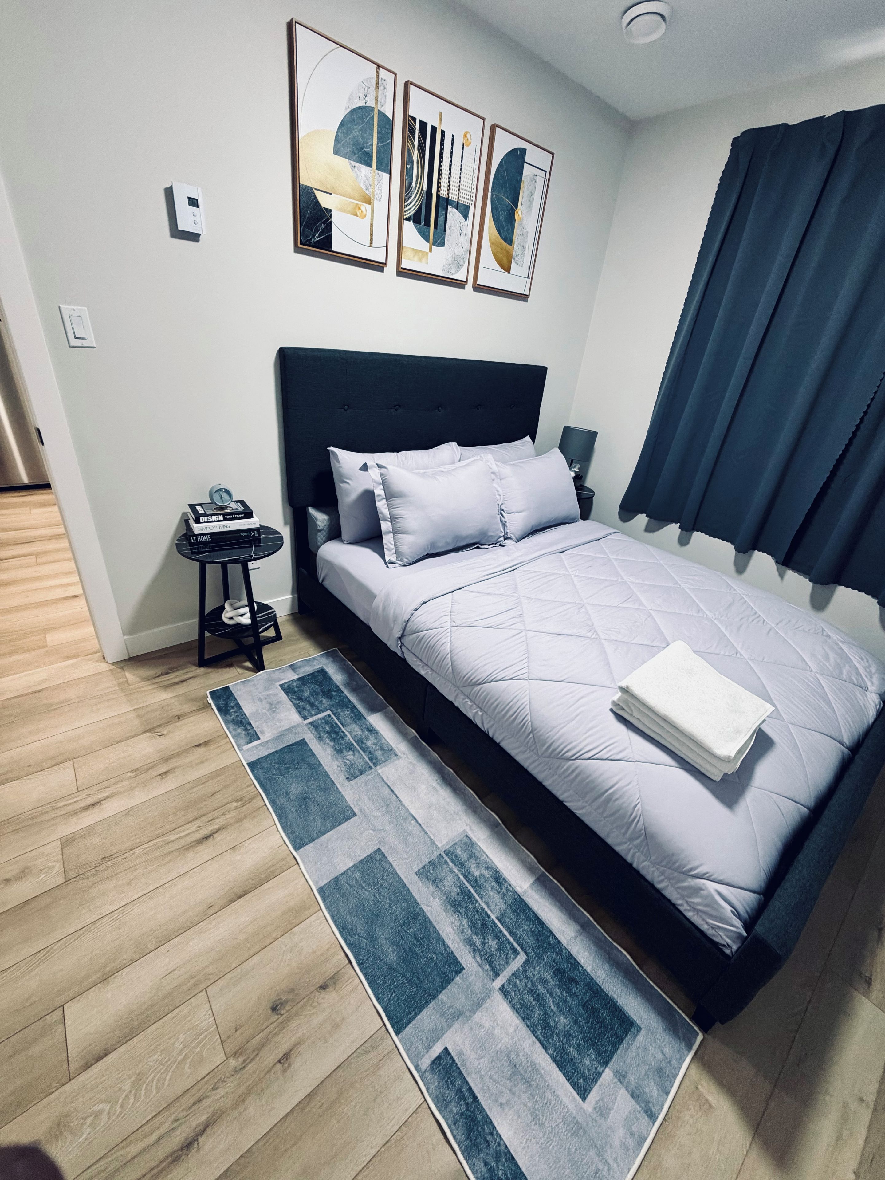 A modern bedroom with a bed, a nightstand, a wall-mounted thermostat, and a geometric-patterned rug.