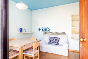 Une pièce comprenant une table à manger en bois, des chaises, un canapé blanc orné d’un coussin à motifs, un luminaire suspendu et une étagère fixée au mur sur laquelle se trouve un vase bleu.
