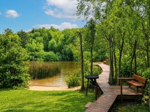 Una terraza de madera con un banco y una mesita, rodeada de abundante vegetación y un lago tranquilo.