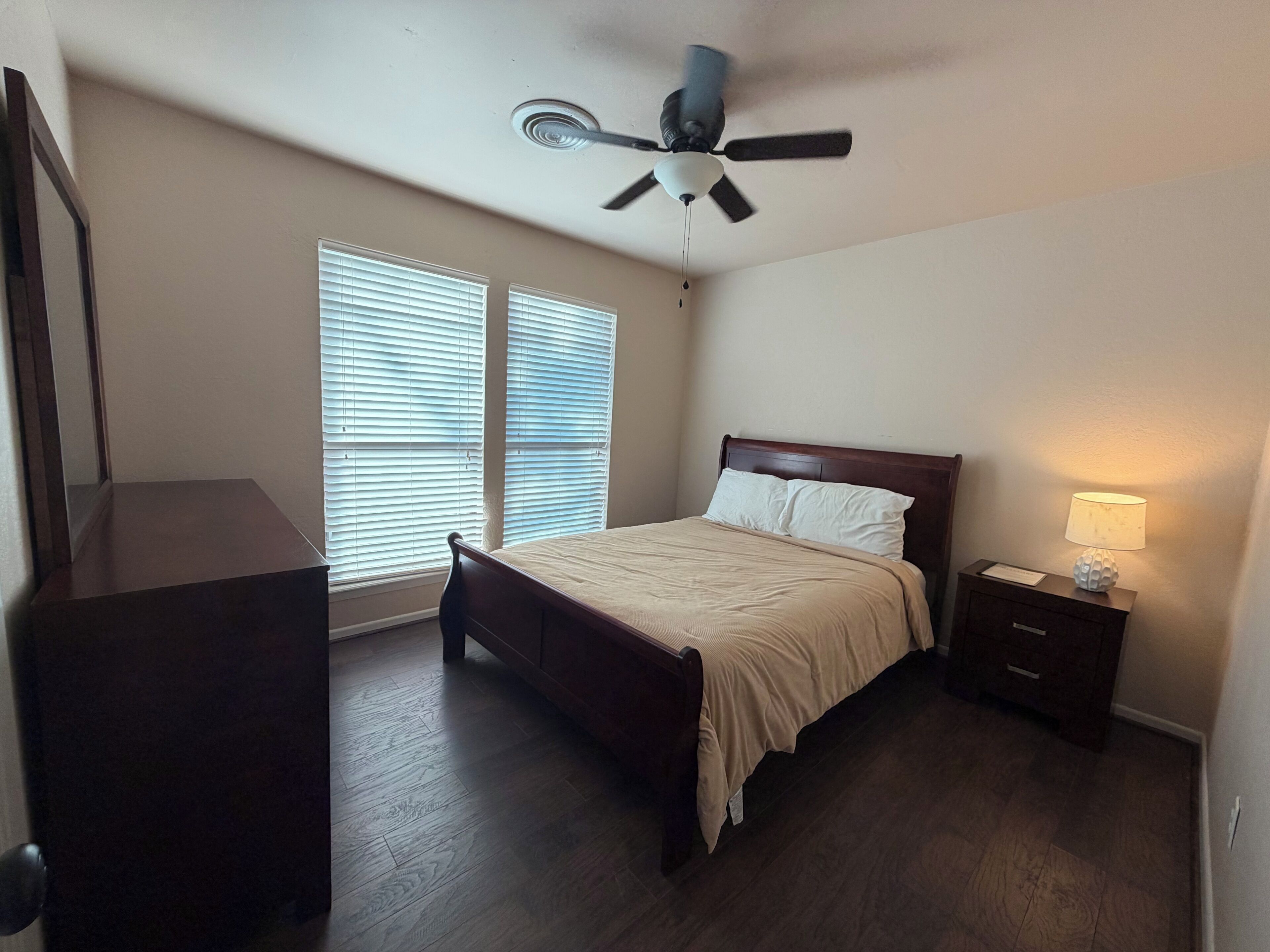 A bedroom with a bed, a nightstand, a lamp, a ceiling fan, and a window with blinds.
