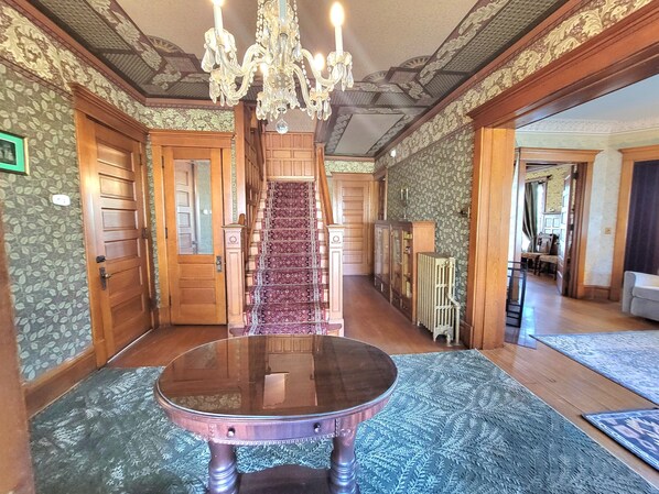Un vaste hall d’entrée avec un escalier en bois, un lustre et un papier peint à motifs.