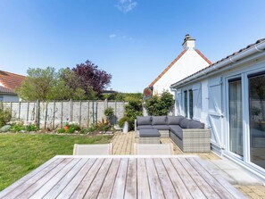 A wooden deck with a sofa and a table, a white building with a red roof, and a wooden fence. - Cosy apartment in villa for 6 people with WIFI, TV and terrace (De Haan)