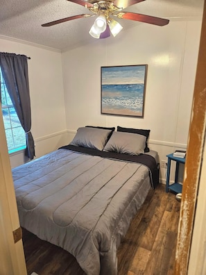 A bedroom with a bed, a ceiling fan, a painting, and a small table. - Sunburst RV Resort (Milton)
