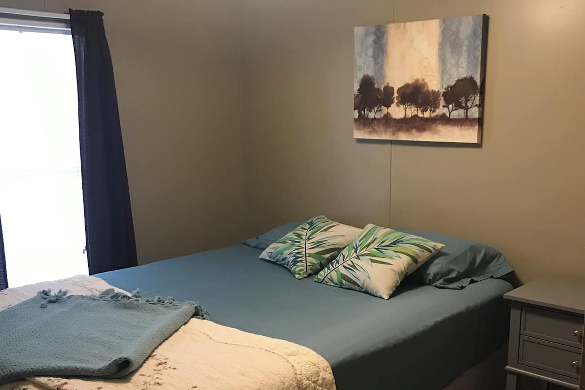 A bedroom with a bed, a nightstand, a painting, and a window with curtains.