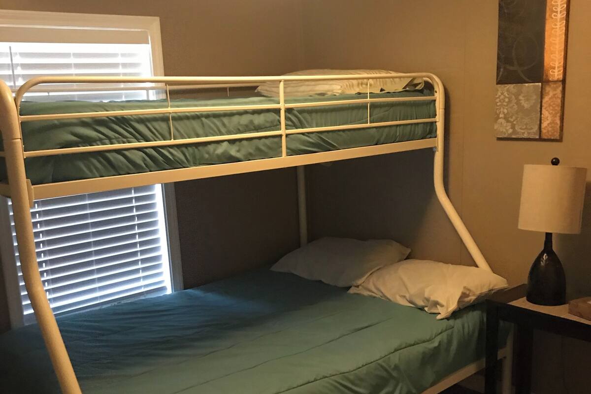 A bunk bed with a green mattress, a bedside table with a lamp, and a window with blinds.